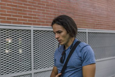 Mavi tişörtlü genç adam kulaklıklarıyla müzik dinlerken binanın yanında dalgın dalgın yürüyor.