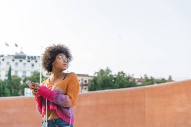 Endişeli kadın, bir yaz günü şehir caddesinde akıllı telefonunu tutarken yan tarafa bakar.