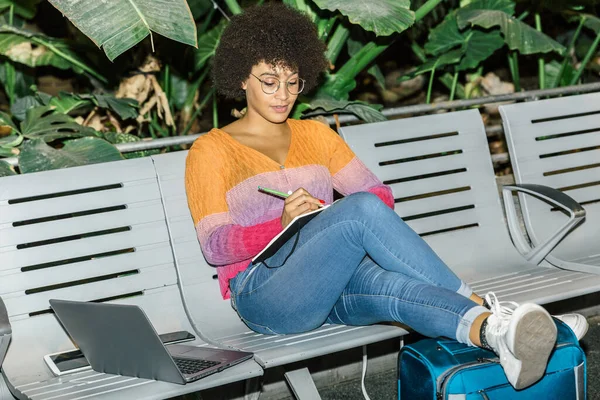 Beautiful mixed race woman writes in her diary while waiting with her luggage sitting on a bench in a station