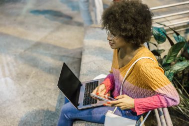 Renkli elbiseli, dizüstü bilgisayarlı, siyah saçlı bir kadının istasyonda oturuşunun yan görüntüsü.