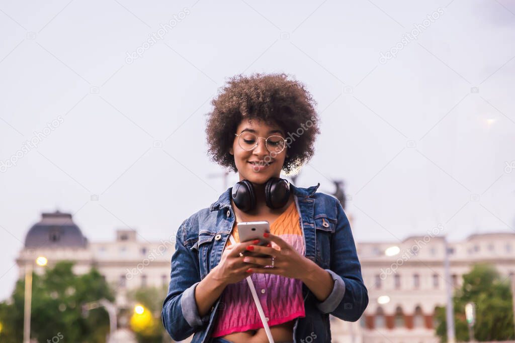 Young dark skinned woman with black headphones around her neck smiles on the street looking at her smartphone