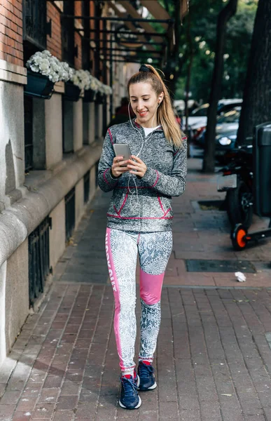 Happy female runner walks through the city before running looking at her smartphone and listening to music