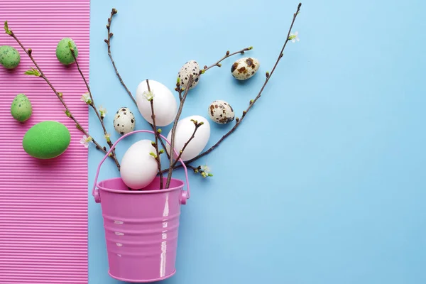 Vue de dessus des œufs de Pâques avec des brindilles vert printemps et seau rose décoratif sur un fond rose-bleu pastel . — Photo