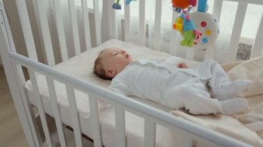 Şirin bebek çocuk büyük pencere yakınındaki beyaz ahşap beşik içinde rahatlatıcı. Bebeğin yüzünde kamera yakınlaştırır