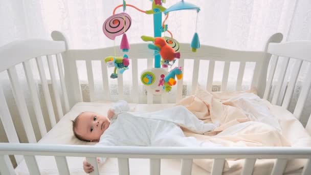 Lindo bebé niño acostado en una cama de madera blanca y jugando con un juguete móvil — Vídeo de stock