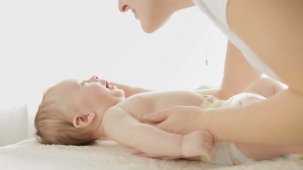 Closeup tiro de jovem mãe falando e beijando seu filho bebê deitado na cama na frente de uma grande janela — Vídeo de Stock