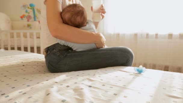 クローズ アップ ショットの母彼女の赤ちゃんとベッドの上に座ってミルクのボトルを取り、赤ちゃんの息子にそれを与えて — ストック動画