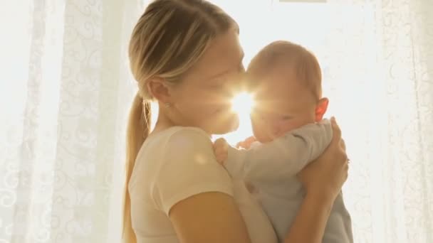 Retrato de la alegre madre joven con su bebé en los rayos del sol delante de la ventana grande — Vídeos de Stock