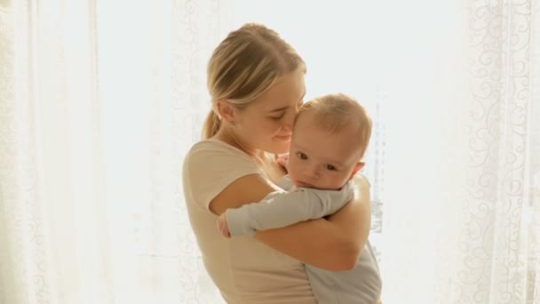 Tonica colpo caldo di felice giovane madre tenendo il suo bambino di 3 mesi alla grande finestra in raggi di sole luminosi — Video Stock