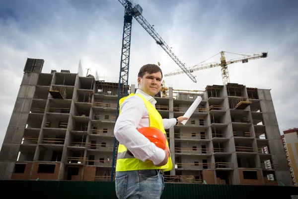 Young engineer in safety vest with blueprints at building site — Stock Photo, Image