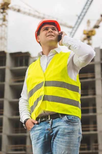 Young smiling engineer in hardhat and safety vest talking by pho — Stock Photo, Image