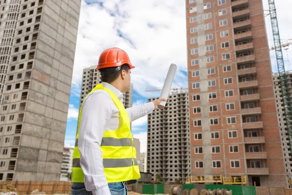 Architect in hardhat pointing with rolled blueprints at building — Stock Photo, Image