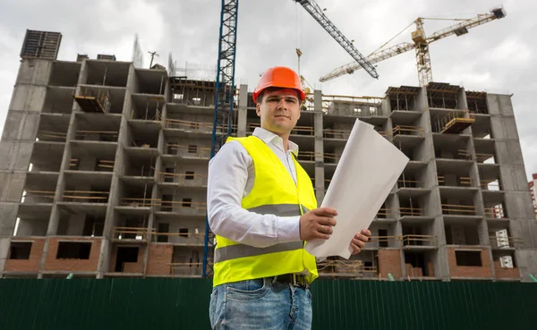 Young smiling engineer on building site checking plans and bluep — Stock Photo, Image