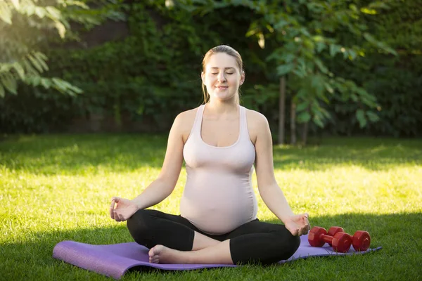 Lotus poz çimenlerin üzerinde oturan güzel gülümseyen hamile kadın — Stok fotoğraf