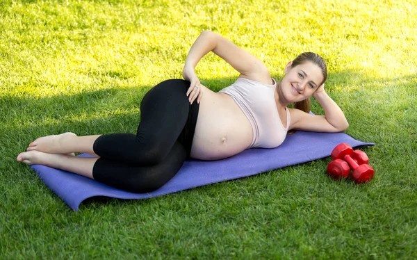 O femeie gravidă zâmbitoare care face exerciții pe saltea de fitness în parc — Fotografie, imagine de stoc
