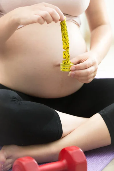 Gravid kvinna poserar med mätning av tejp och hantlar på gym — Stockfoto