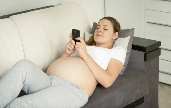 Jovem grávida deitada no sofá e usando smartphone — Fotografia de Stock