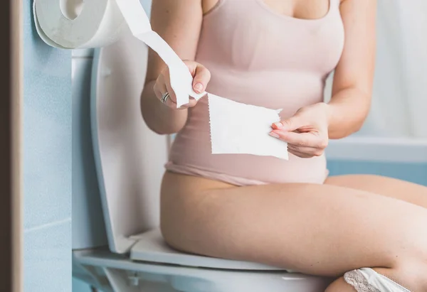 Young woman sitting on toilet and tearing off paper tissue — Stock Photo, Image