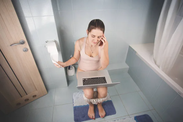 Mujer sentada en el inodoro y usando el portátil y hablando por teléfono —  Fotos de Stock