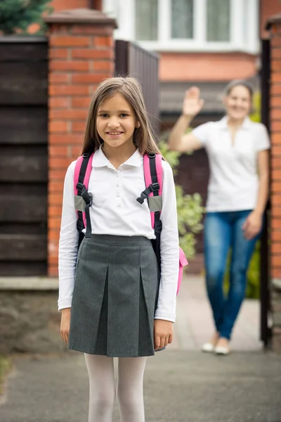 Portrait of cute schoolgirl standing in front of house. Mother w