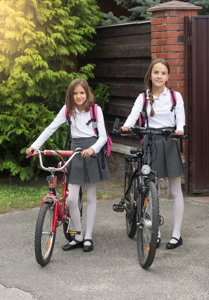 Две улыбающиеся девочки в школьной форме прогуливаются с велосипедами по улице — стоковое фото