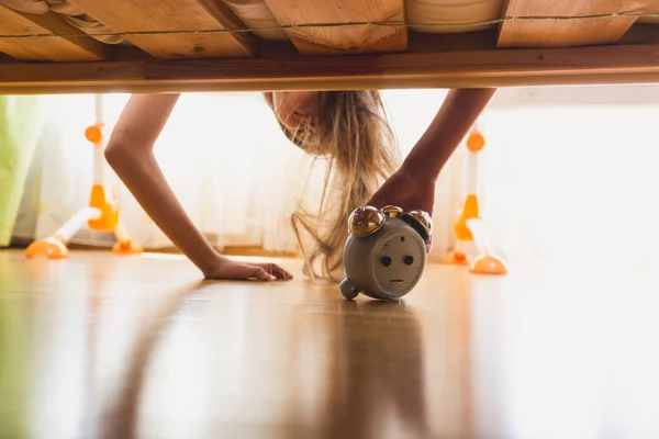 Annoyed girl reaching for alarm clock under the bed at early mor — Stock Photo, Image