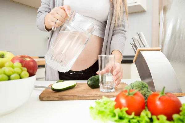 Νεαρή έγκυος γυναίκα ρίχνει νερό σε ποτήρι στην κουζίνα — Φωτογραφία Αρχείου
