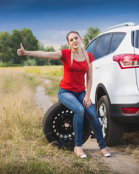 Young sad woman sitting on wheel at broken car and hitchhiking — Stock Photo, Image