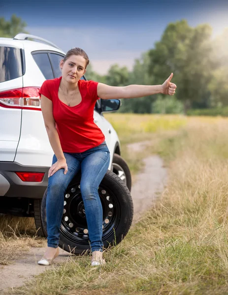 Jeune femme bouleversée assise à une voiture cassée et en auto-stop — Photo