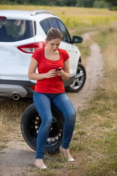 Young upset woman siting on spare tire next to broken car at fie — Stock Photo, Image