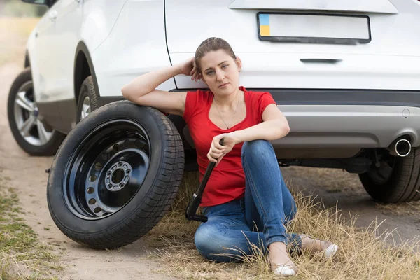 Молодая расстроенная женщина сидит на земле, опираясь на сломанную машину — стоковое фото