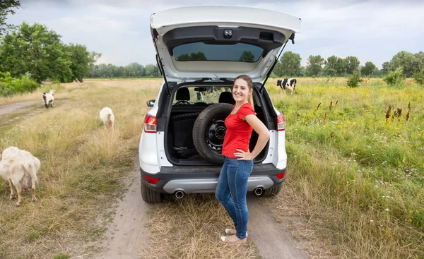 Junge Frau steht an kaputtem Auto auf Feld neben Ziegenherde — Stockfoto