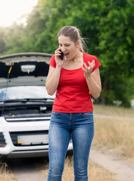 Angry young woman standing at broken car and talking by mobile p — Stock Photo, Image