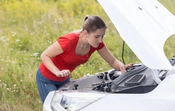 Joven hermosa mujer mirando debajo de la capucha de coche roto en fi — Foto de Stock