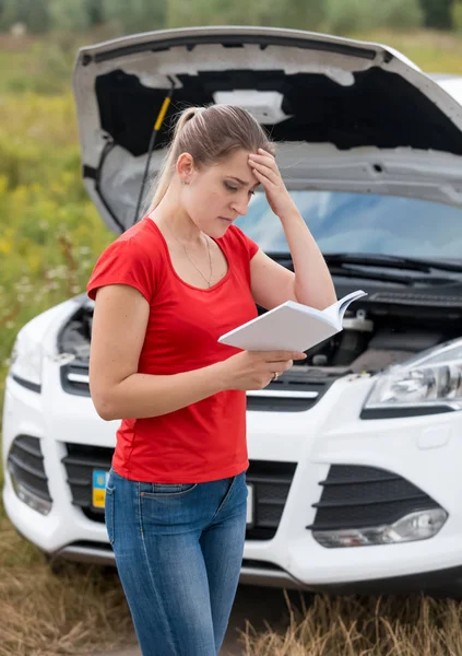 Portrait of young woman reading owner manual at broken car — Stock Photo, Image