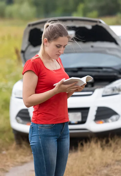 Smutná mladá žena čte návod k obsluze na rozbité auto v louce — Stock fotografie