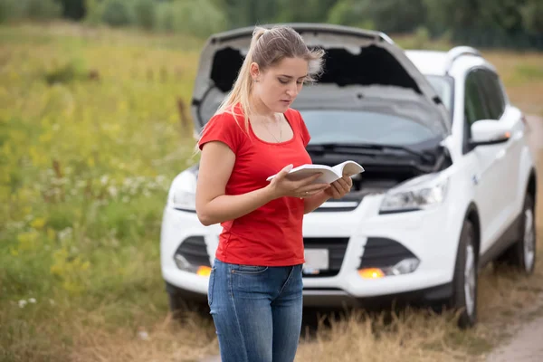 Depressed woman looking at her broken car and reading owner manu — Stock Photo, Image