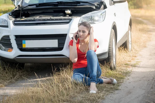 Portrait of young woman sitting next to broken car and calling f