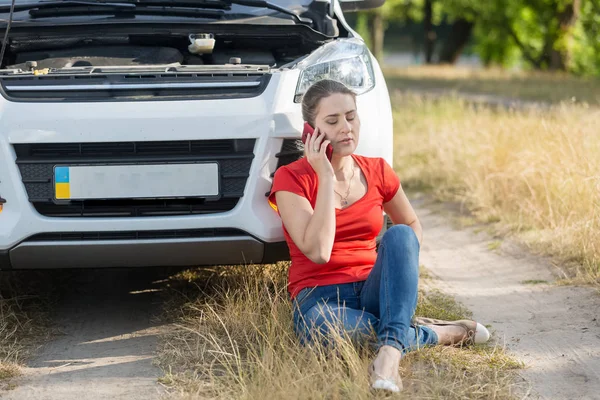 Young female driver sitting on ground next to broken car and cal — Stock Photo, Image