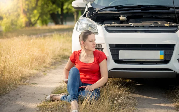 Female driver sitting on ground next to broken car and calling f — Stock Photo, Image