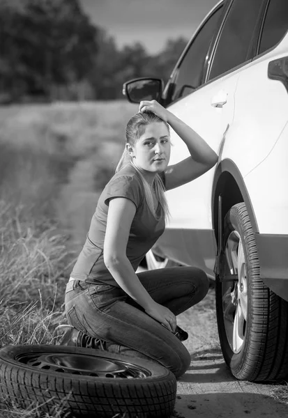 Immagine in bianco e nero di sconvolto giovane donna cambiando ruota auto su — Foto Stock