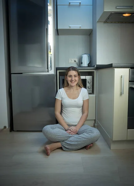 Женщина в пижаме сидит на полу на кухне рядом с открытым холодильником — стоковое фото