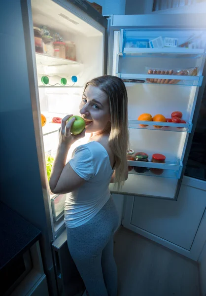Молода жінка в піжамі їсть піцу у відкритому холодильнику вночі — стокове фото