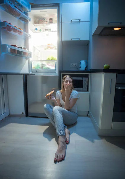 Wanita yang duduk di lantai dapur di samping kulkas terbuka dan makan — Stok Foto