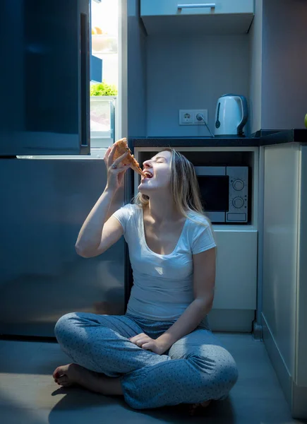 Голодная женщина ест пиццу на полу кухни поздно вечером — стоковое фото