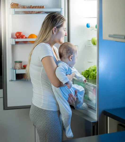 Mutter hält Baby-Sohn in der Hand und schaut in Kühlschrank — Stockfoto