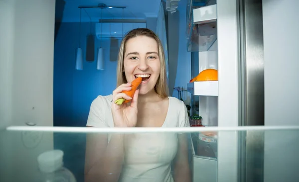 Feliz joven mujer tomando zanahoria del refrigerador y comiéndola —  Fotos de Stock