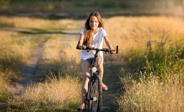 Chica sonriente cabalgando en bicicleta en los prados al atardecer — Foto de Stock