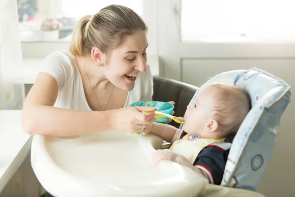 Jovem mãe sorrindo dando molho de maçã para seu bebê em cadeira alta — Fotografia de Stock