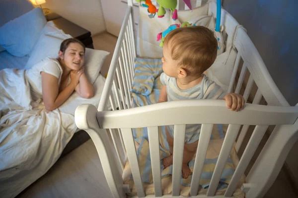 Cute baby standing in crib and looking at tired mother that fell — Stock Photo, Image